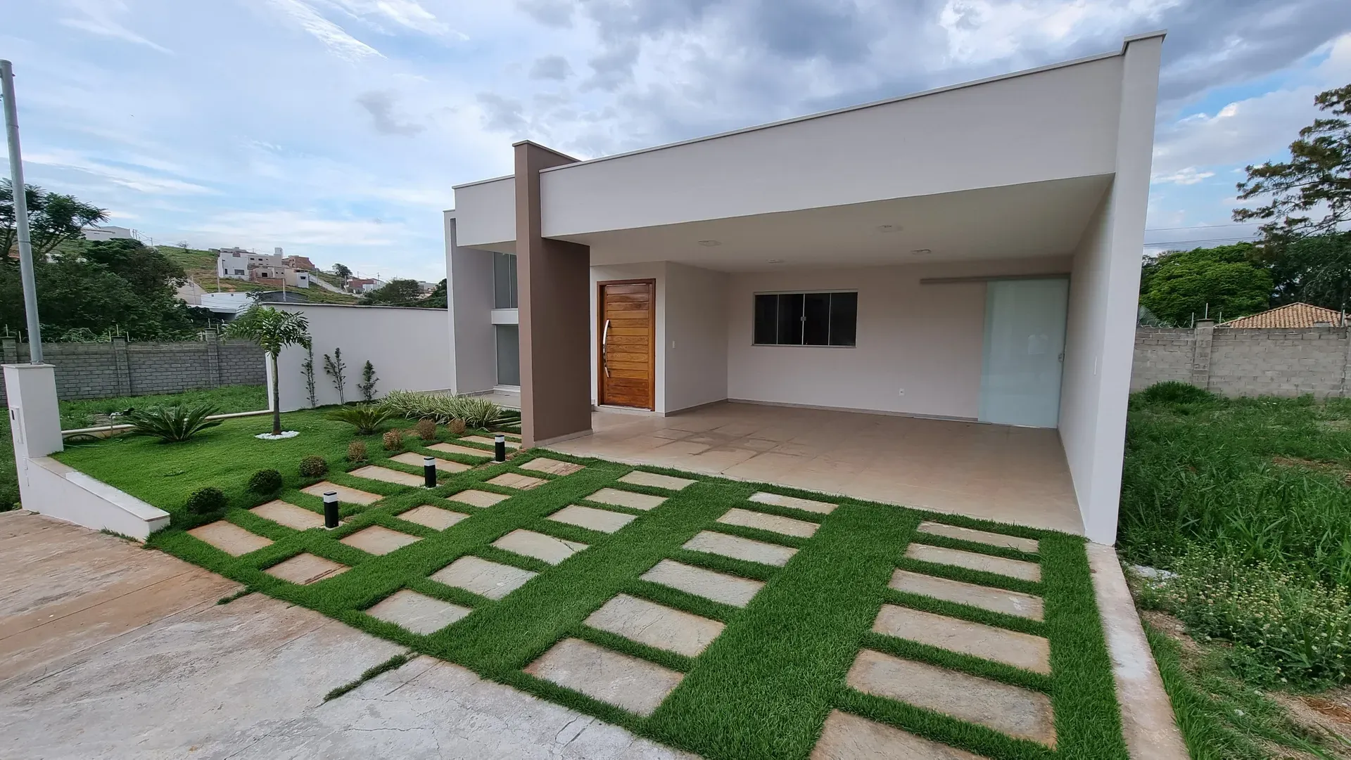 Fachada da Casa do Gran Ville com garagem coberta, jardim de grama e pedras e a porta de entrada em madeira