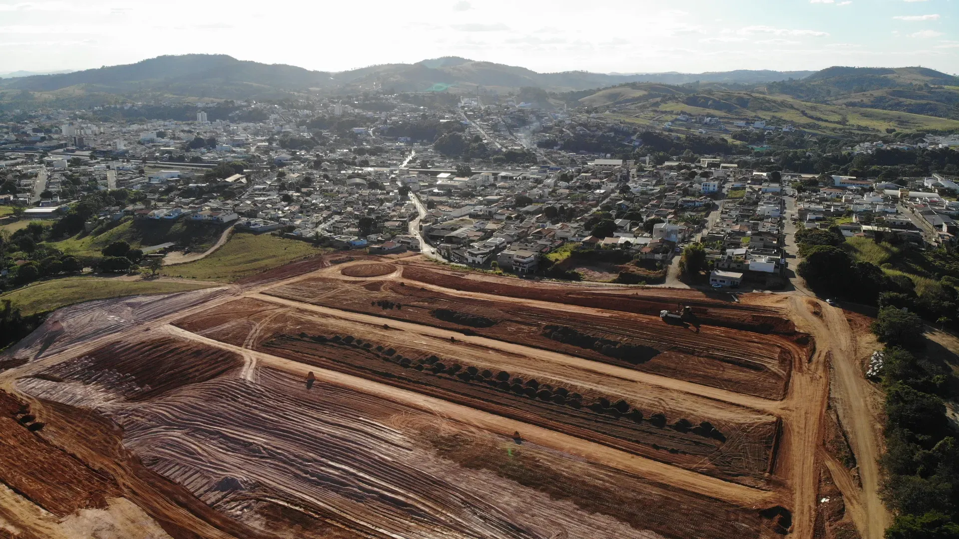 Vista aérea do Portal da Serra em obras ao entardecer, com os platôs de terraplenagem em primeiro plano e a cidade de Perdões ao fundo