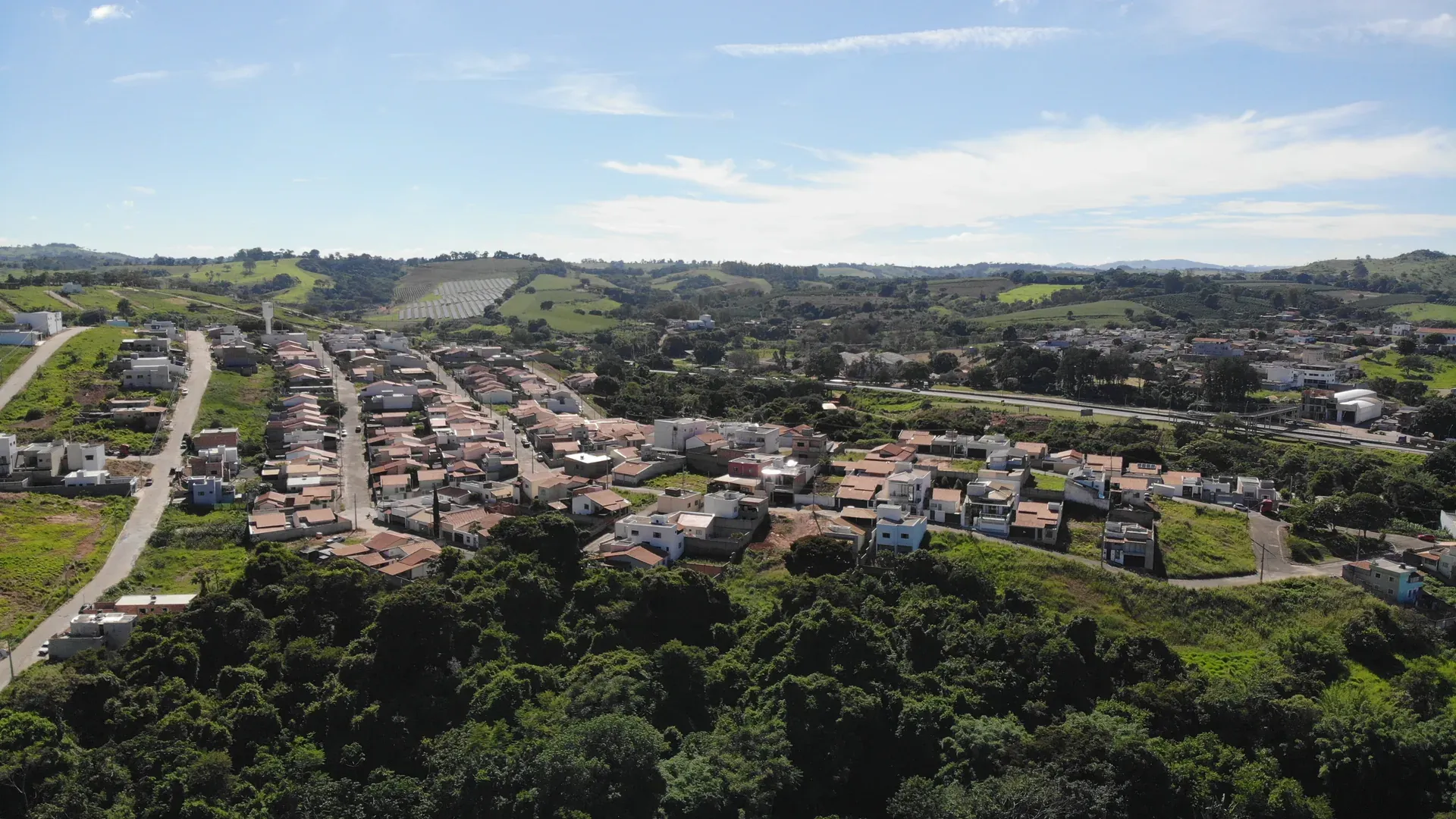 Vista aérea ampla do Jardim Centenário em direção às colinas do Sul de Minas, com mata nativa em primeiro plano