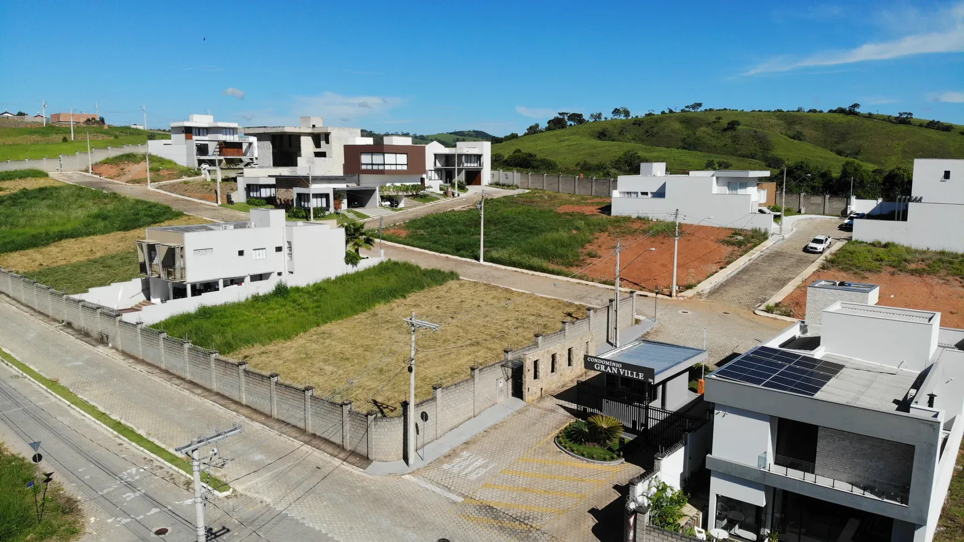 Vista aérea da portaria principal do Condomínio Gran Ville com identificação visível, casa moderna de alto padrão com placas solares ao lado e residências ao fundo