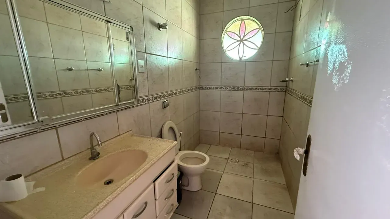 Banheiro da Casa do Lourdes com bancada e cuba, espelho amplo, área de chuveiro e janela redonda em vitral