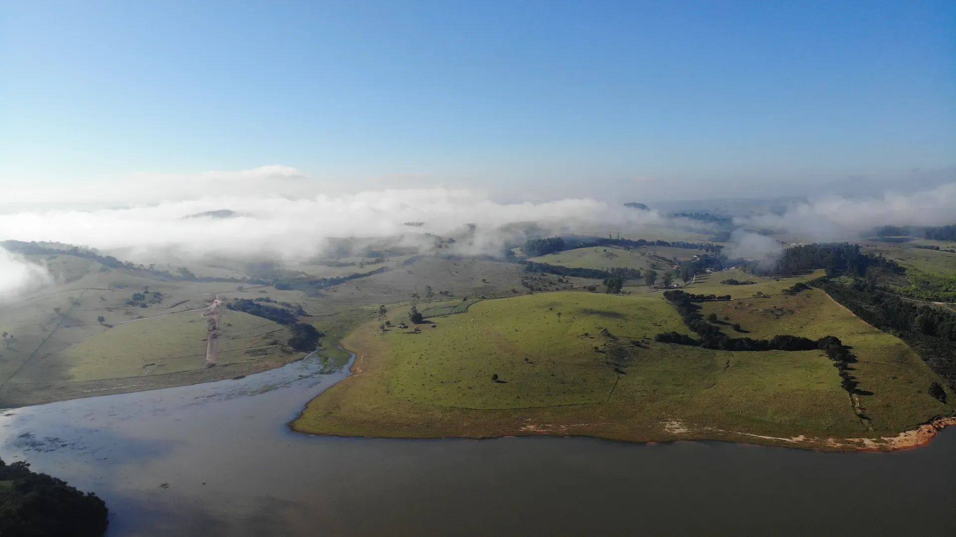Margem do lago e pastagens da fazenda, com neblina baixa cobrindo os morros ao fundo