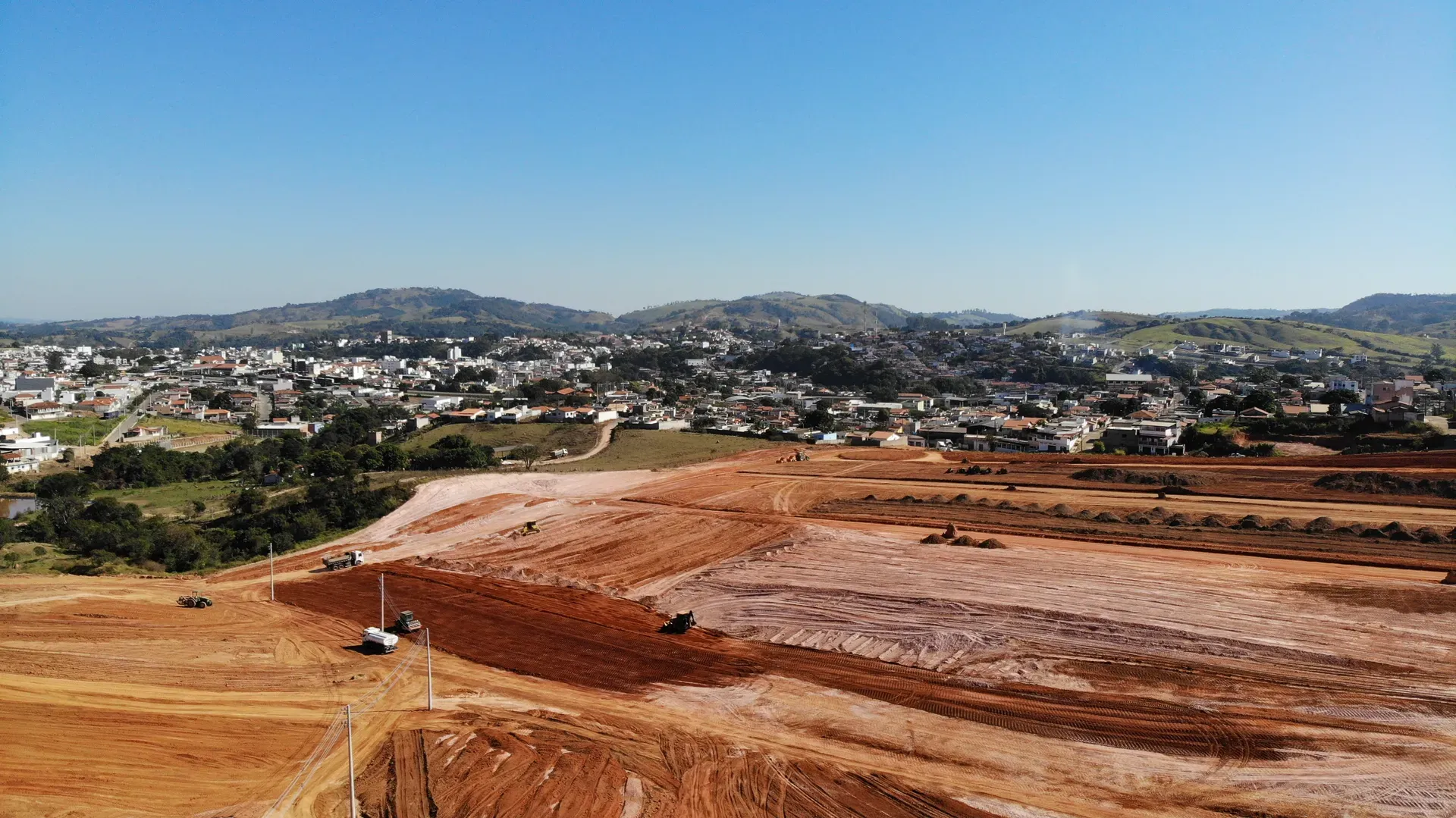 Aérea das frentes de terraplenagem do Portal da Serra sob céu azul, com maquinário e a cidade ao lado