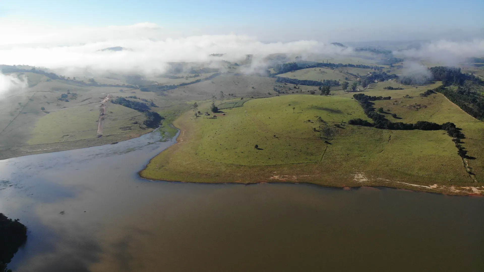 Pasto à beira d’água da Fazenda Coelhos, com a água do lago e a neblina sobre a serra