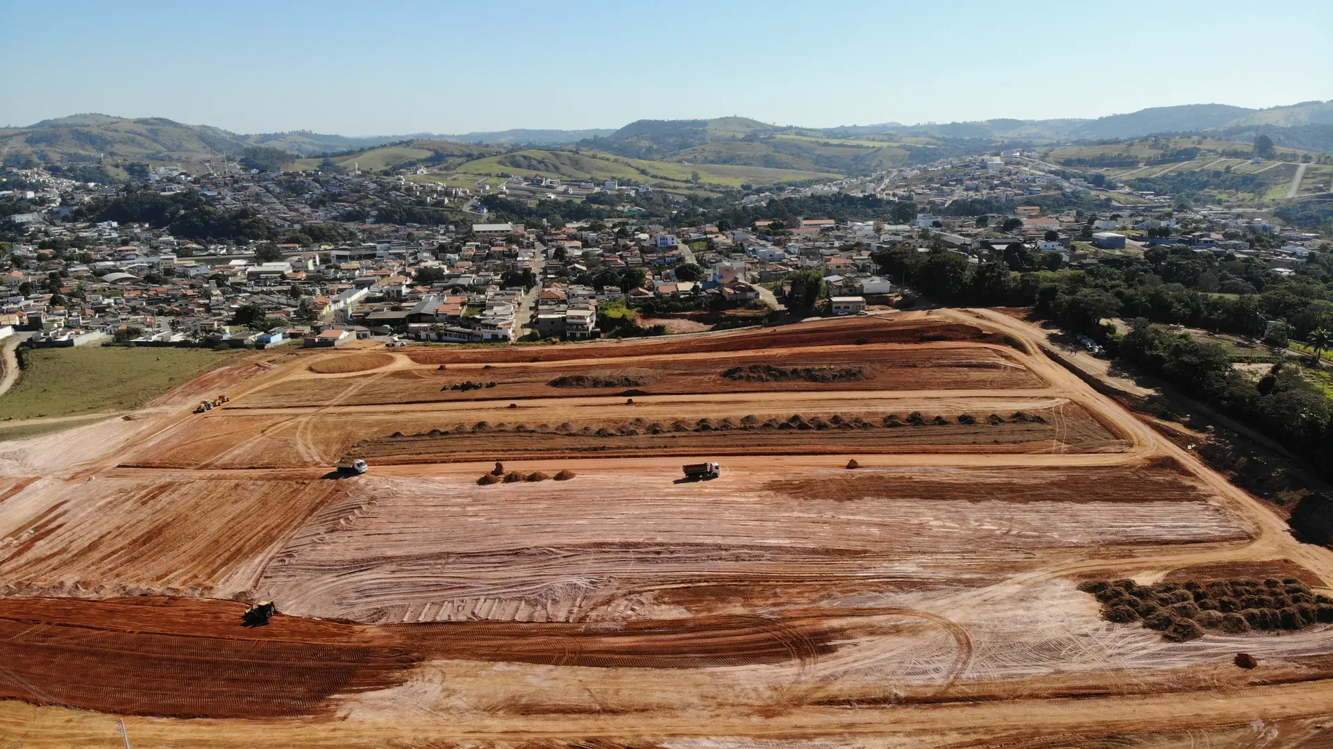 Aérea dos platôs do Portal da Serra em terraplenagem ao fim da tarde, com maquinário e a cidade de Perdões ao fundo