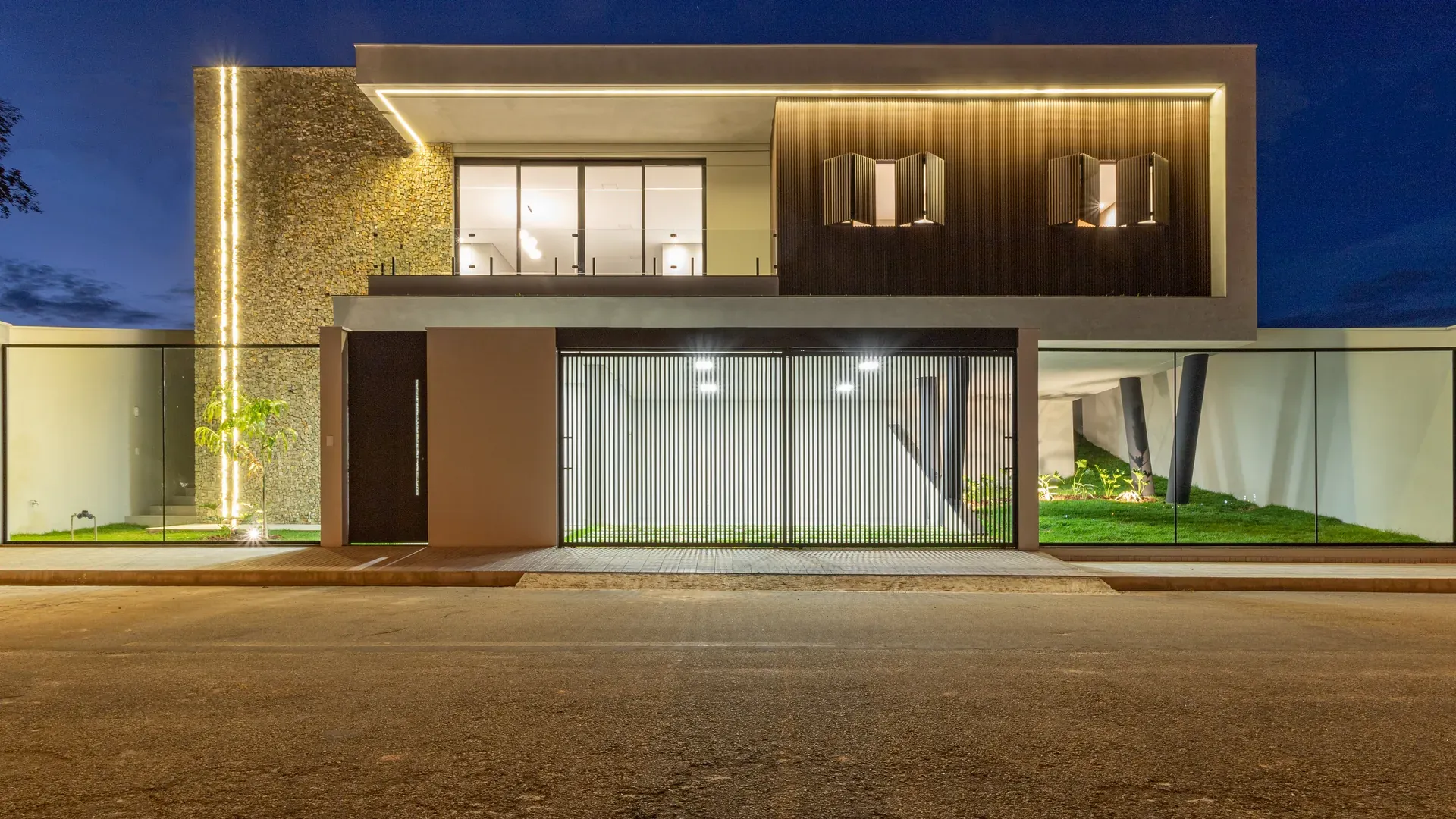 Fachada da Casa Monte Verde ao anoitecer, sobrado moderno iluminado em Perdões/MG, com pavimento térreo envidraçado e o céu azul do fim de tarde