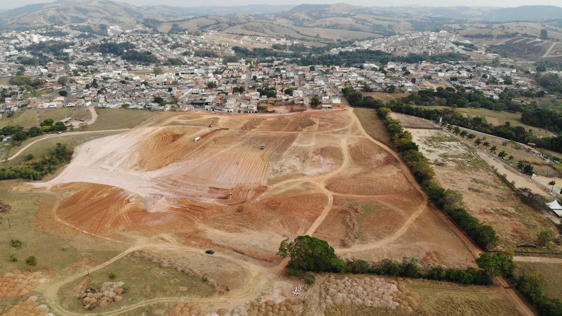 Vista aérea do Residencial Portal da Serra em obras em Perdões/MG, com a topografia em platôs do loteamento, a cidade ao lado e a serra ao fundo