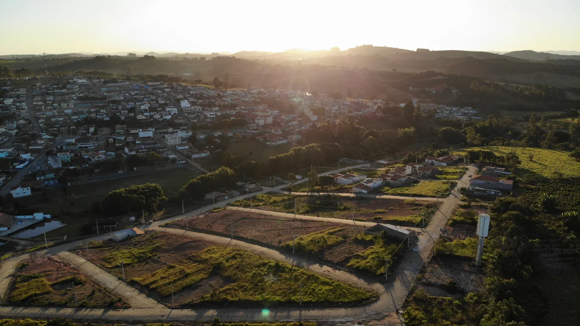 Vista aérea ao pôr do sol de São Sebastião da Estrela/MG com o Residencial Bela Vista à frente, ruas demarcadas e o distrito iluminado pela luz dourada do fim de tarde