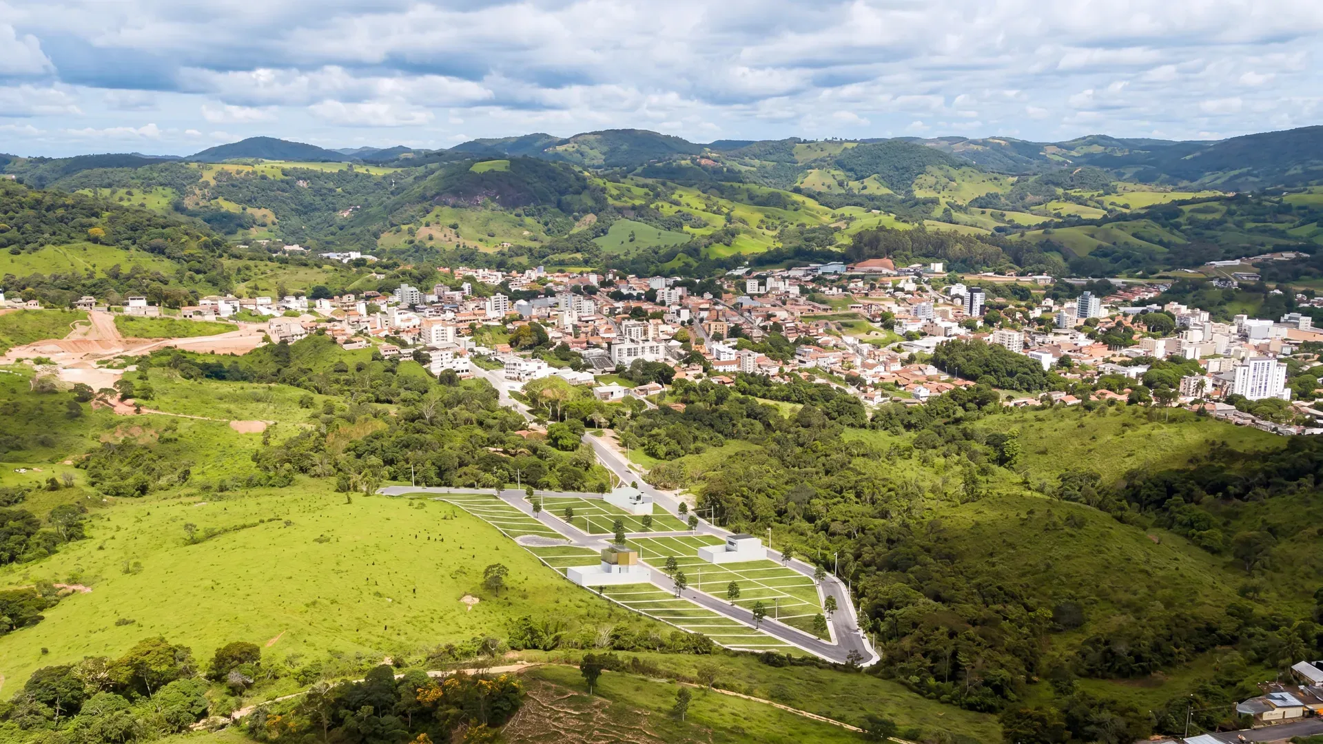 Vista aérea do Residencial Veredas da Serra em Itaguara/MG, com os lotes e as vias do loteamento no primeiro plano verde, a cidade de Itaguara ao centro e a serra do Sul de Minas ao fundo