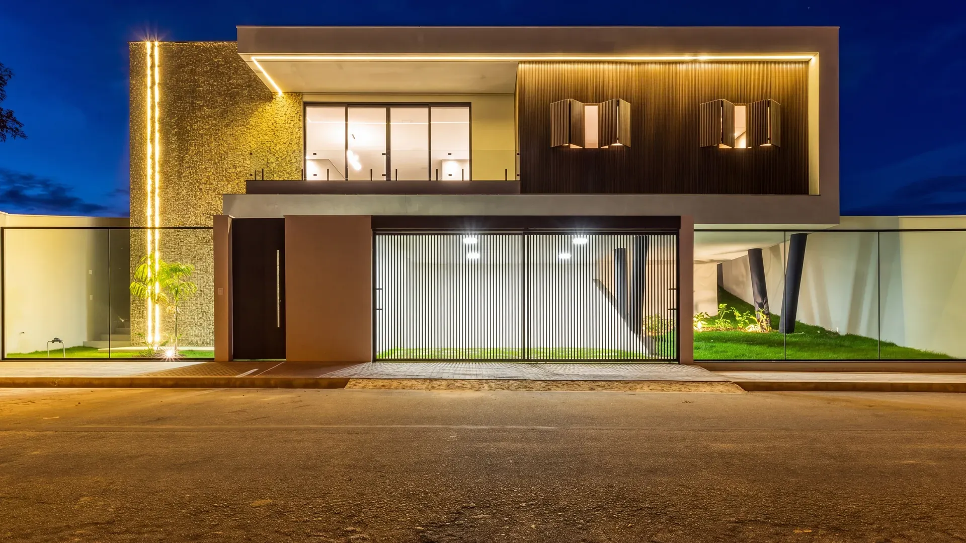 Fachada da Casa Mirone ao anoitecer, sobrado moderno iluminado em Perdões/MG, com pavimento térreo envidraçado e o céu azul do fim de tarde