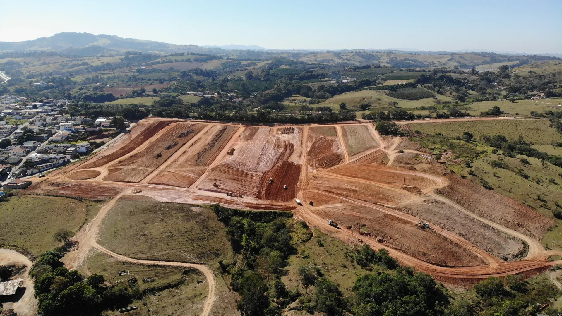 Vista aérea do Portal da Serra em obras em Perdões/MG, com a topografia em platôs do loteamento em terraplenagem, a cidade ao lado e a serra ao fundo