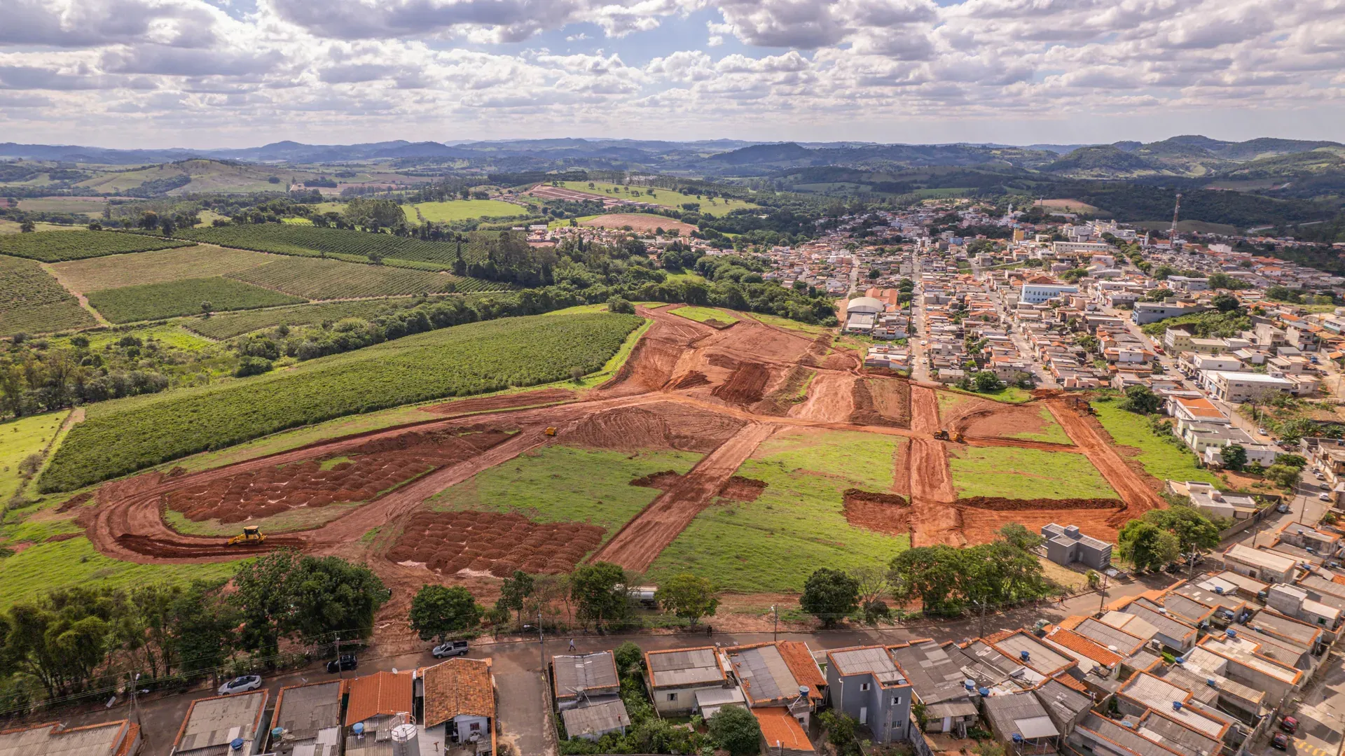 Vista aérea do Residencial Ouro Verde em obras em Cana Verde/MG, primeiro bairro planejado da cidade, com os lotes em terraplenagem e a cidade e a serra ao fundo