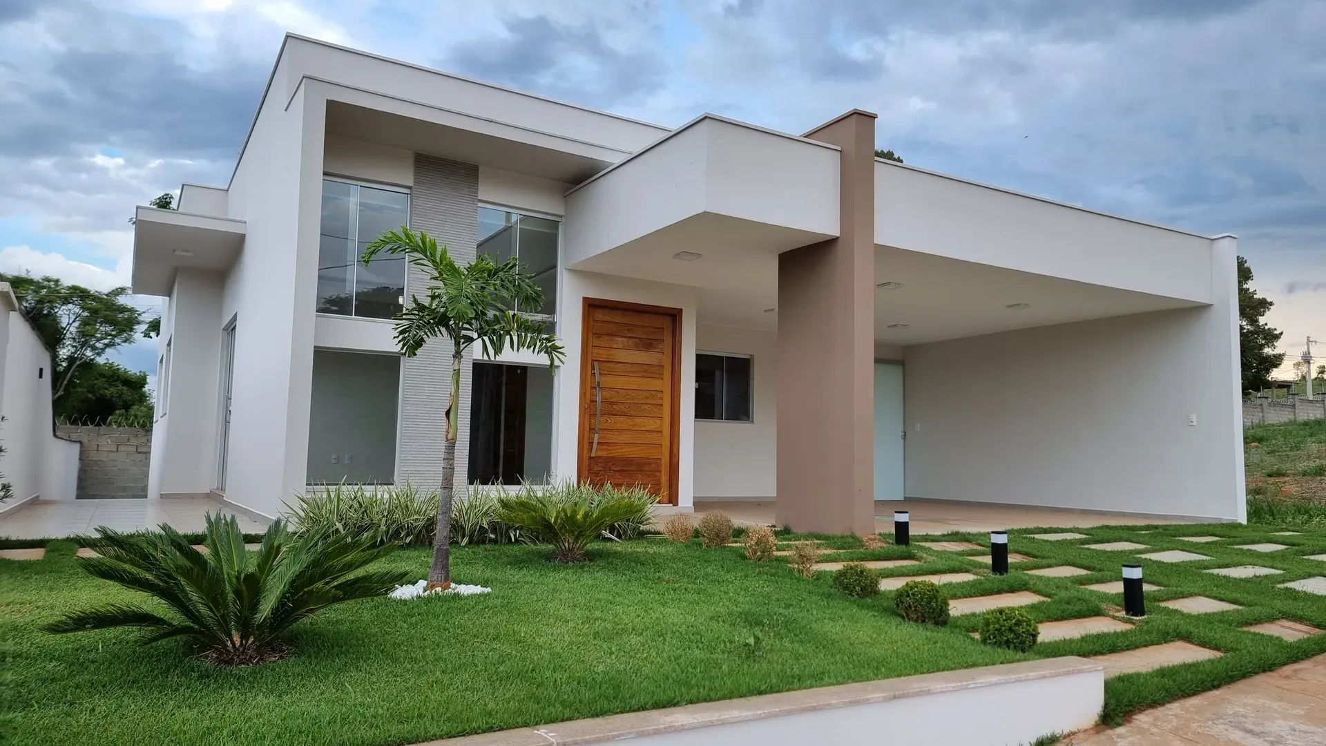 Fachada frontal da Casa do Gran Ville, residência moderna térrea em Perdões/MG, com porta pivotante de madeira, amplo jardim e garagem coberta