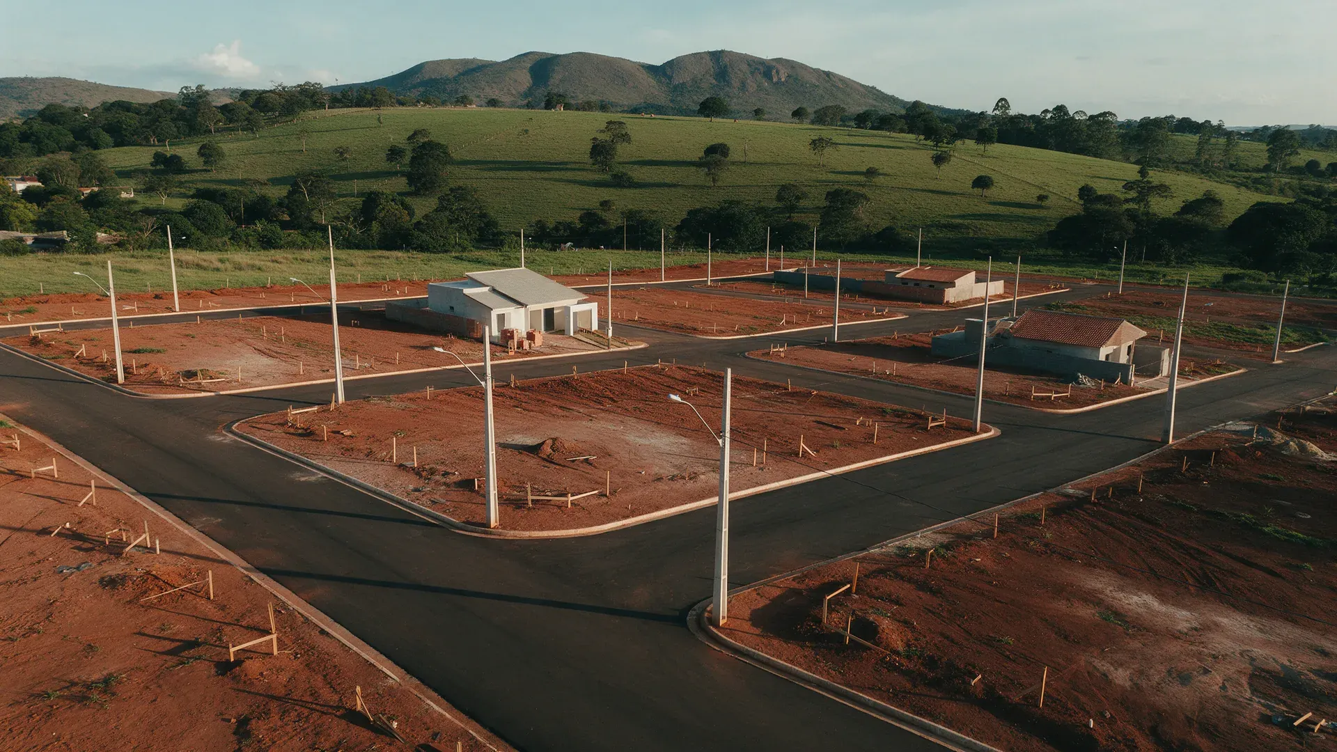 Vista aérea de um loteamento recém-urbanizado na borda de uma cidade do Sul de Minas, com ruas asfaltadas, lotes demarcados, casas em construção e serra ao fundo no fim da tarde