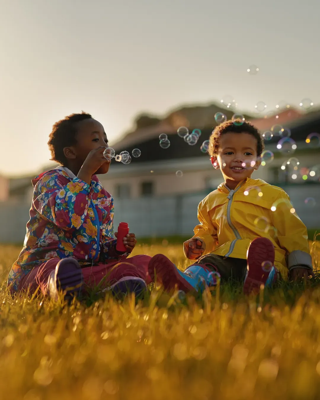 Duas crianças brincam com bolhas de sabão num gramado ao entardecer, com as casas do bairro ao fundo