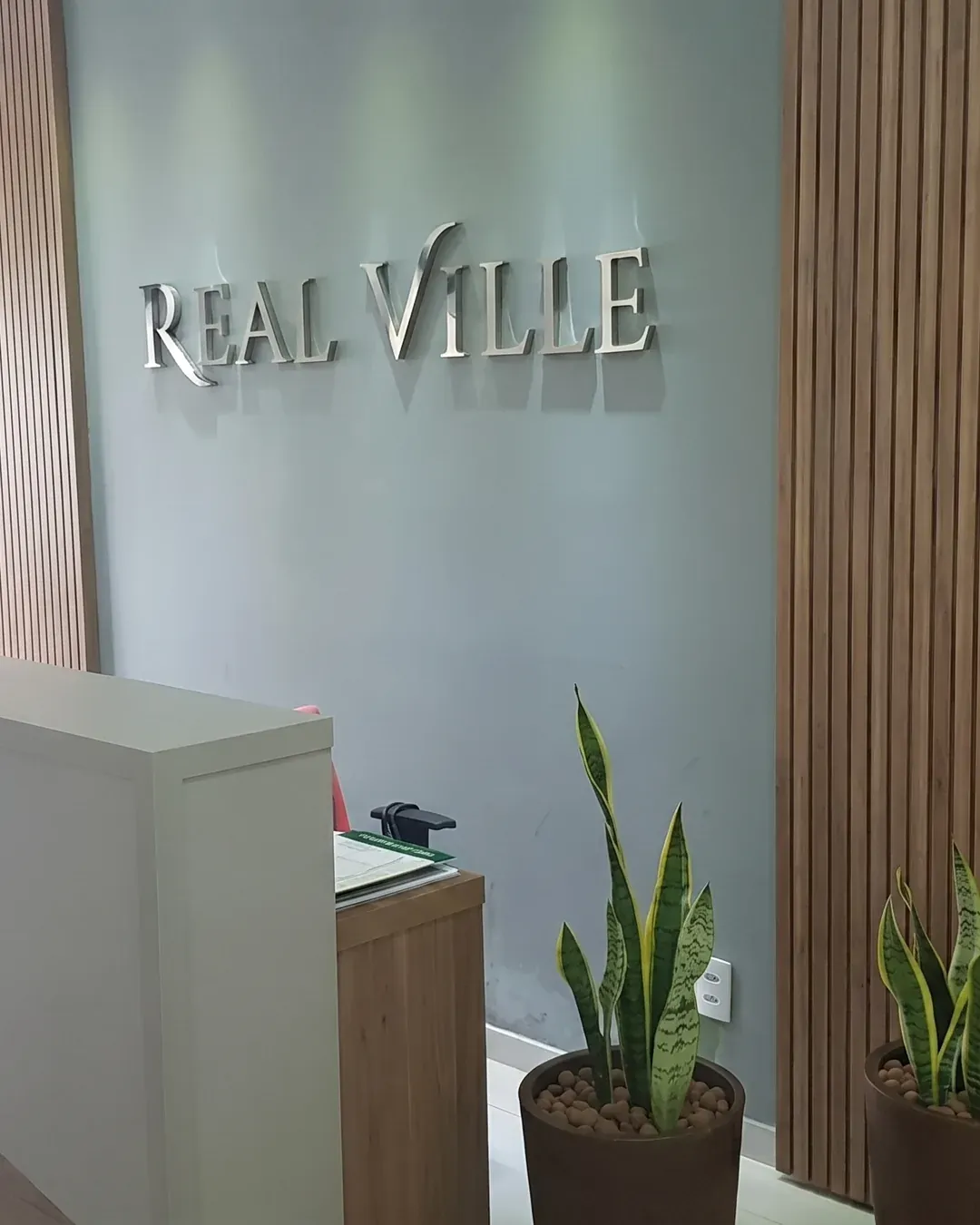 Recepção do escritório da Real Ville Urbanismo em Perdões/MG, com o letreiro REAL VILLE na parede, balcão de atendimento e plantas ornamentais.
